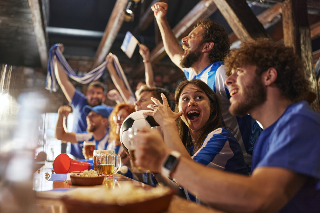 Group of cheerful sports fans cheering while watching a soccer match on TV in a bar. Focus is on woman with ball.